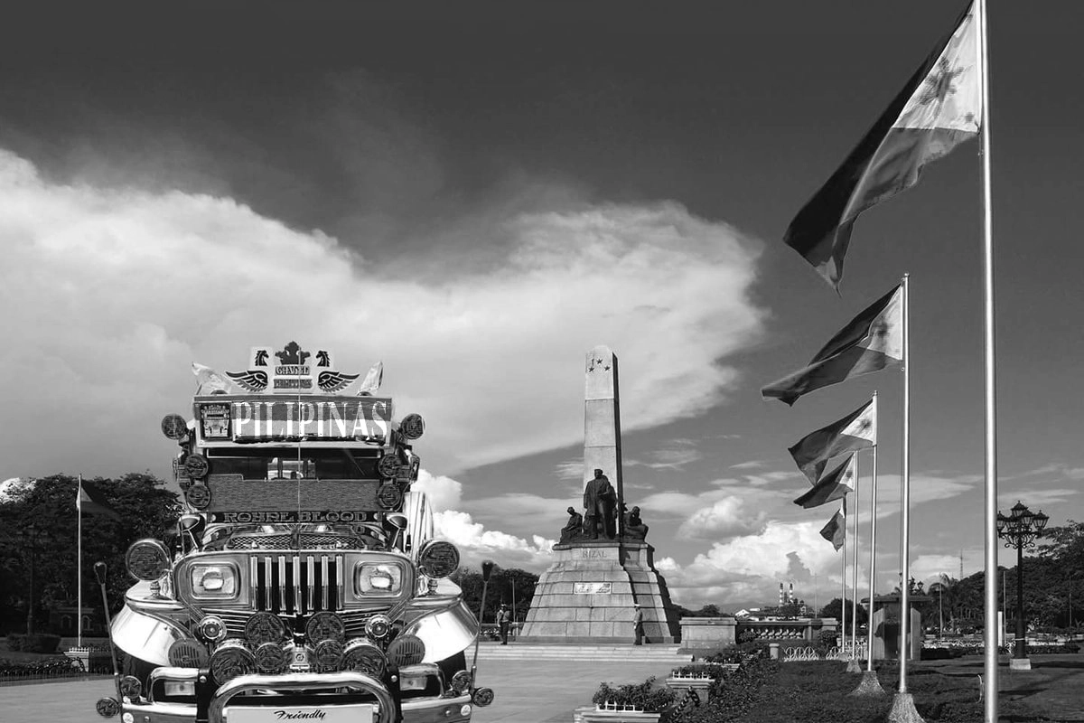 Photo of a jeepney taken in front of the Rizal monument in Luneta park during a sunny day, with poles of Philippine flags waving on the right hand side.